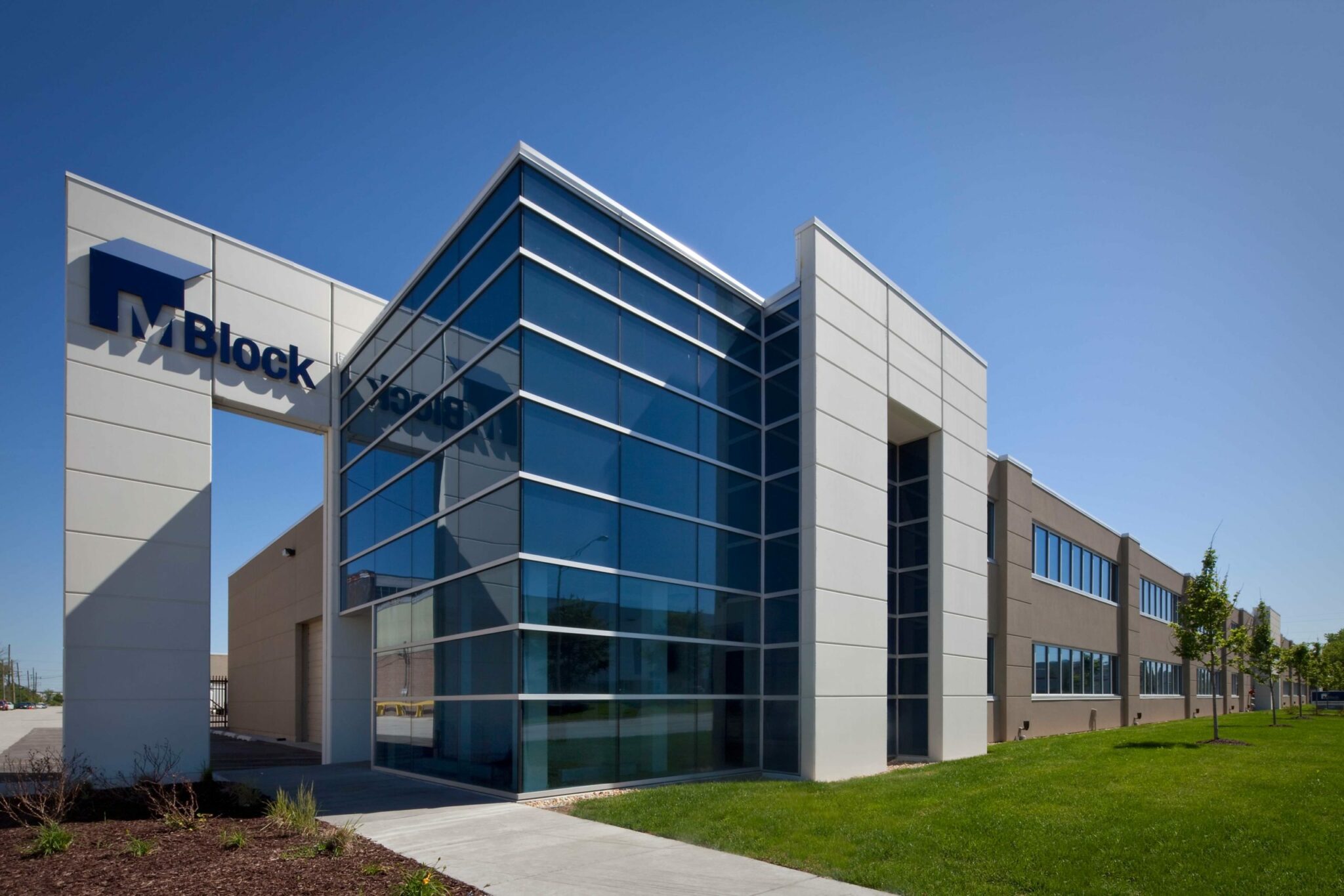 M. Block and Sons' large, new corporate building has floors of windows mirroring the blue sky. A stand-out structure has the words "M. Block" on the top with a sidewalk underneath it. The rest of the building has walls made of brick and other materials that designate the division between corporate offices and warehouse areas.