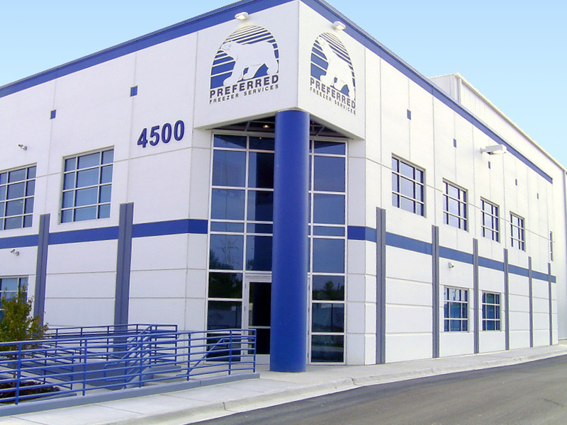 A view of the corner of a large blue and white facility, with mirrored windows at the entrance. The text on both corners of the building say, "Preferred Freezer Services."