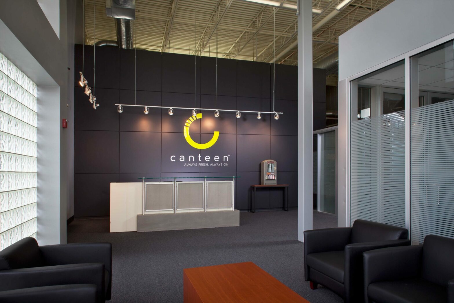 A welcome area with glass walls and comfortable seats. A reception area is in the back, and behind the desk is the word “Canteen” under their logo.