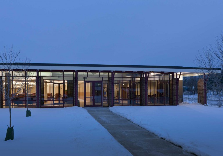 An exterior picture of Montini Catholic High School’s one-story building renovated by Keeley Construction, Inc. The prominent wall is primarily windows, allowing a view of the warm interior light, contrasting with the twilight and white glittering snow outside.