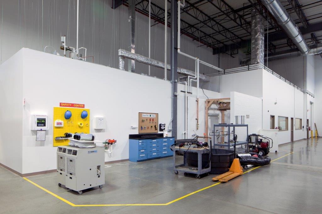 The inside of the new warehouse of National Technical Systems’ building. The large space has workspaces, a corkboard with helmets labeled “Safety Center,” mechanic’s drawers, and an automatic defibrillator on the wall.