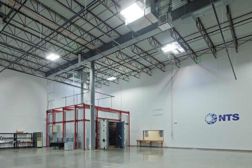 The ceiling of the large warehouse and laboratory has a metal grid, the walls are crossed with pipes and conduits, and the corner holds a large mechanism covered with a red metallic grid. The letters “NTS” are on the wall for National Technical Systems.