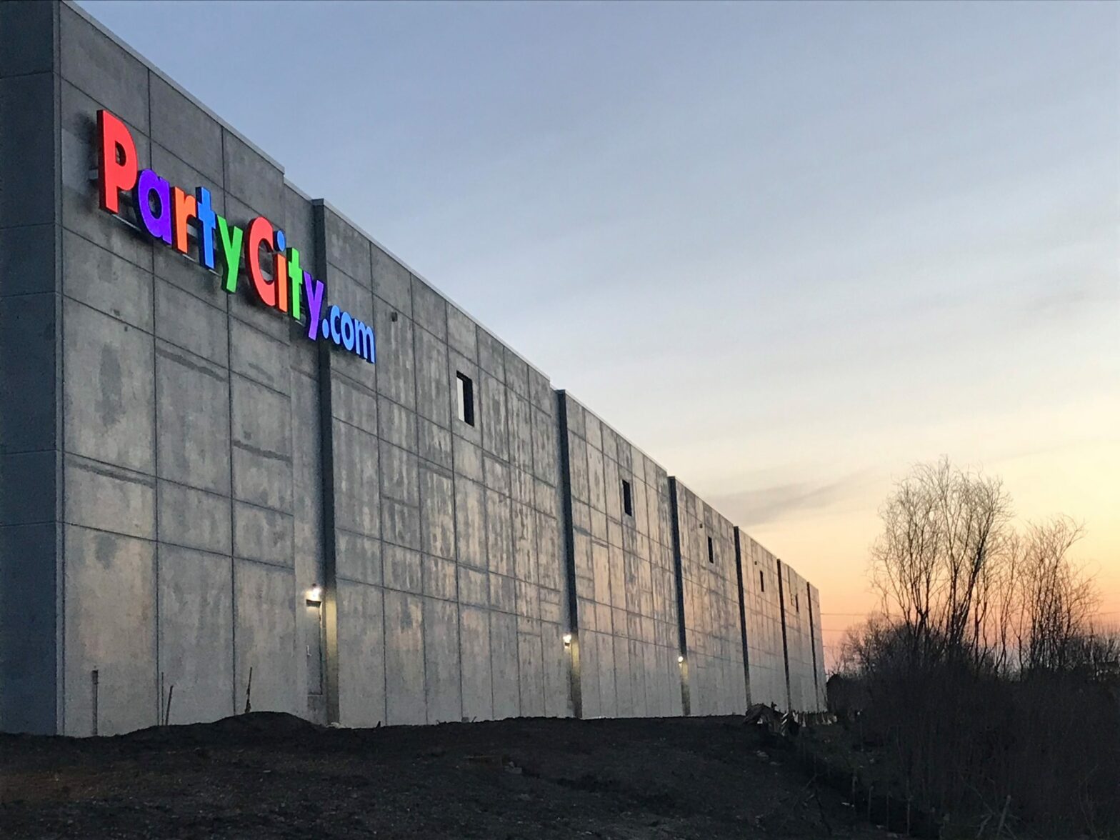 The colorful "Party City" sign illuminates the angled view of the side of the Party City expansion as the sun rises behind the winter trees.