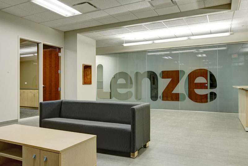 A reception area contains a couch and a desk in front of a frosted glass wall. The word “Lenze” in large letters is not frosted and easily readable.