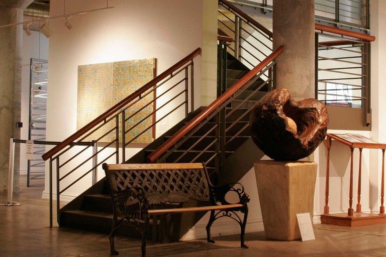 A wooden park bench sits next to a large circular structure placed to the side of the metal staircase in the lower level gallery space in the National Vietnam Veterans Art Museum. Keeley Construction was instrumental in renovating the 80-year-old damaged building.