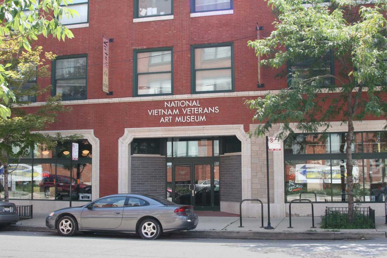 The red-brick exterior and front entrance to a renovated space on a city block. A gray car is parked in front of the building. The sign above the door reads “National Vietnam Veterans Art Museum.”