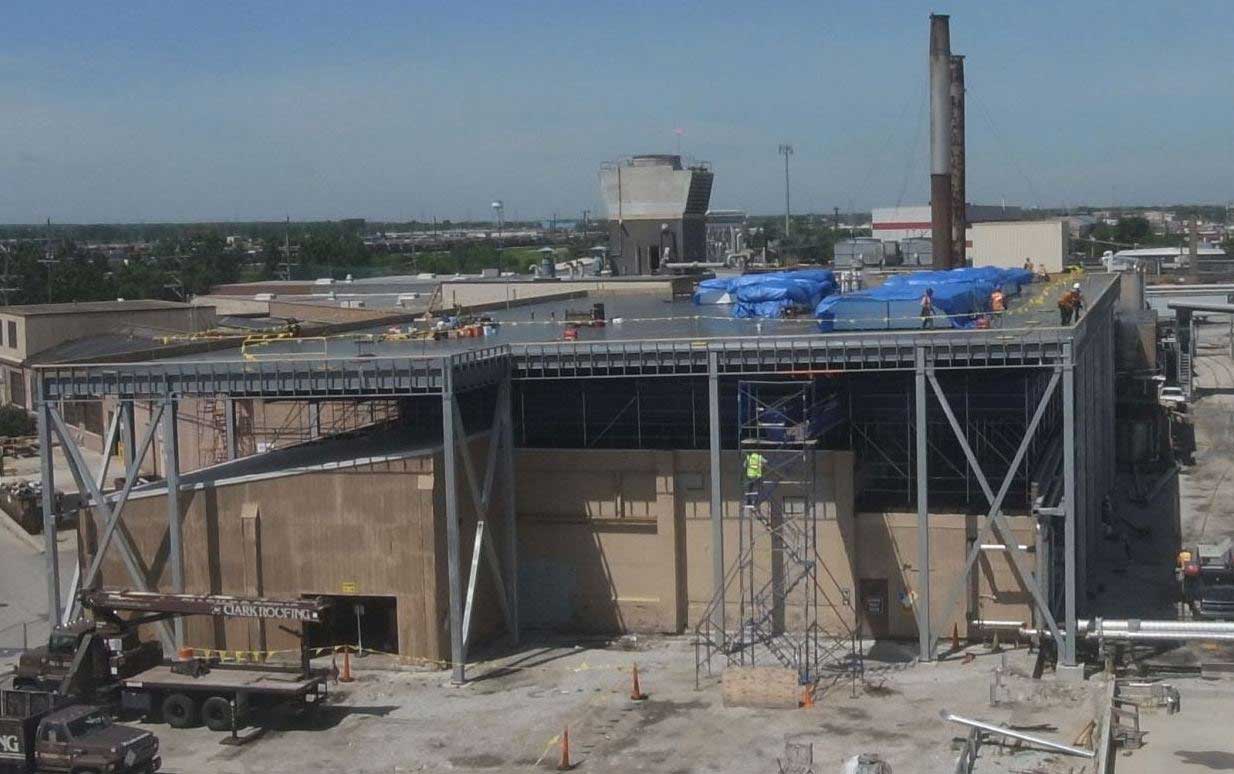 A large new roof structure is built over the existing Nalco Chemical building while construction workers stand on the top and climb the scaffolding. Blue tarp covers supplies on the new roof being built by Keeley Construction, Inc. while the facility was fully operational.