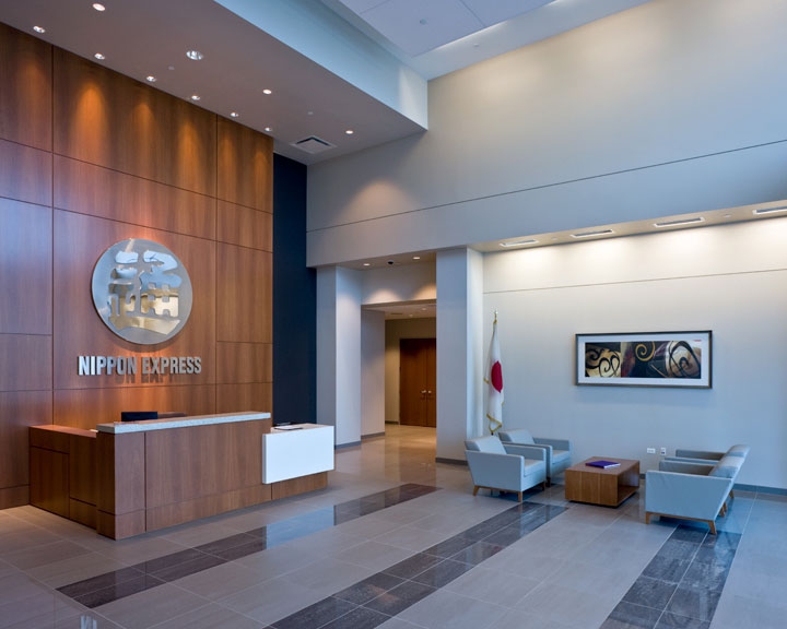 The modern waiting room has contemporary chairs around a small coffee table and a large tan reception desk in front of a wood-paneled wall. The words “Nippon Express” are in silver letters below a circular sign with Japanese characters on the wall behind reception. A Japanese flag is near the waiting room wall.