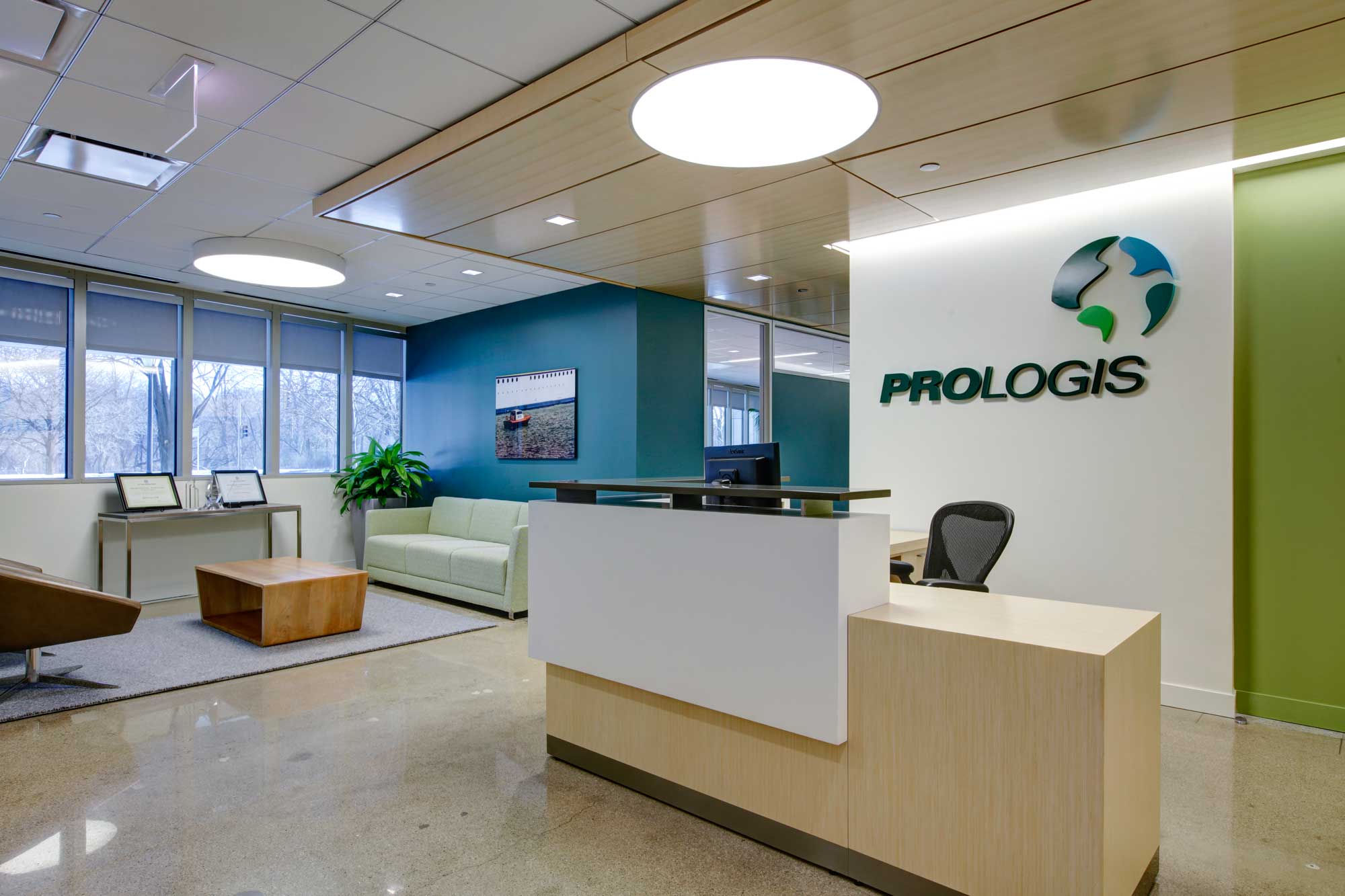 An interior reception area with the words "Prologis” below a swirled logo on the wall shows modern furnishings and fresh blue walls. This O'Hare Industrial Park Façade Renovation by Keeley Construction is a sustainability LEED Gold project.