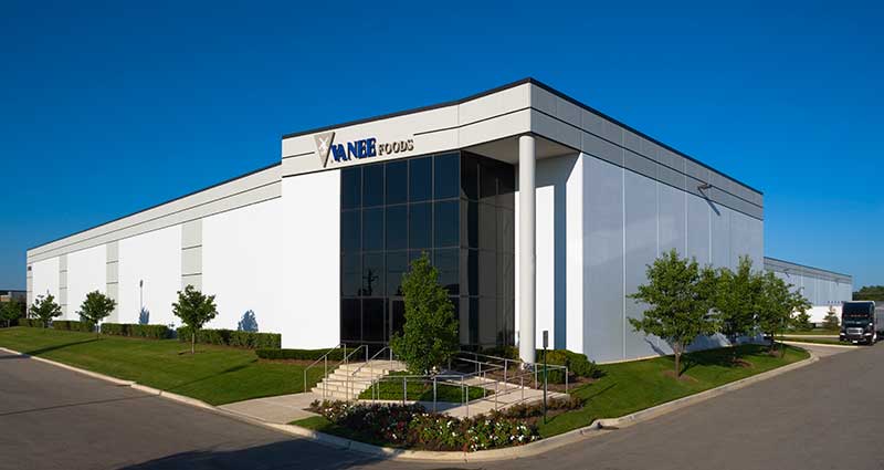 A large, white corporate office and warehouse facility stands against a clear blue sky. Steps and a wheelchair ramp lead to the front door. A logo consisting of a grey triangle and the words “Vanee Foods” are above the windowed front entrance.