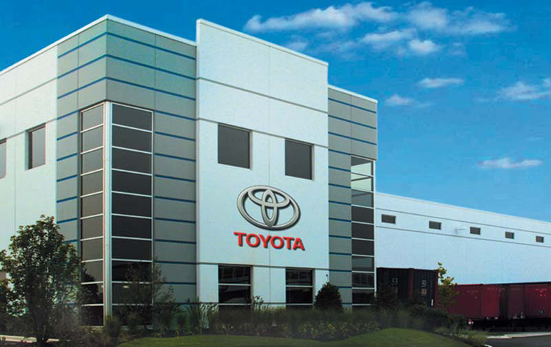 The front of a large office building and warehouse pictured on a bright sunny day is inviting. The words “Toyota” and the Toyota logo hang off the building’s top floor.