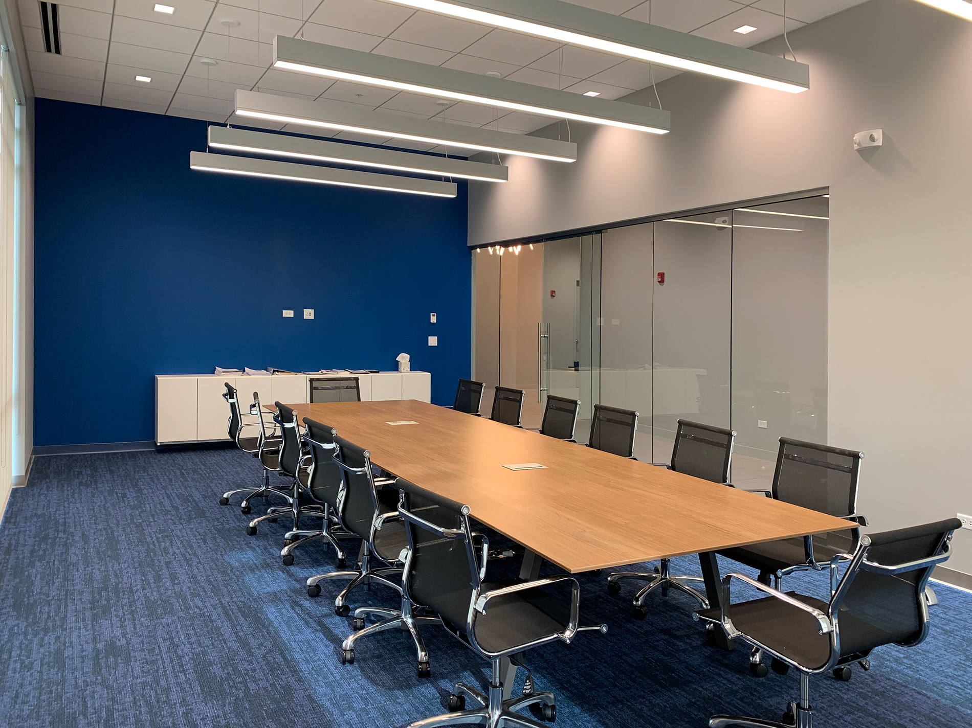 A narrow conference room with a long table has a glass wall on the right and a lot of daylight.