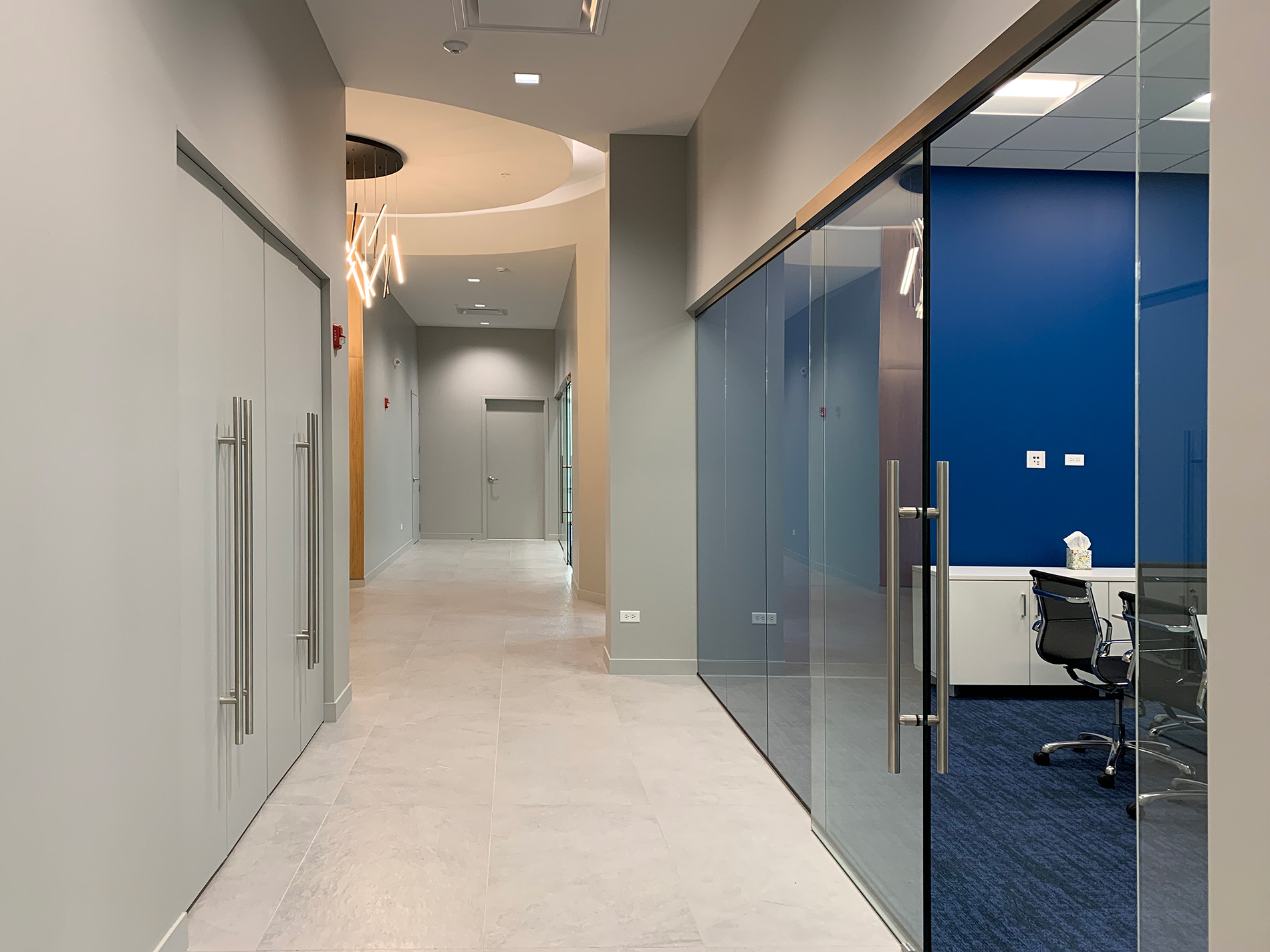 A long hallway with a blue wall and chairs in a room to the right.