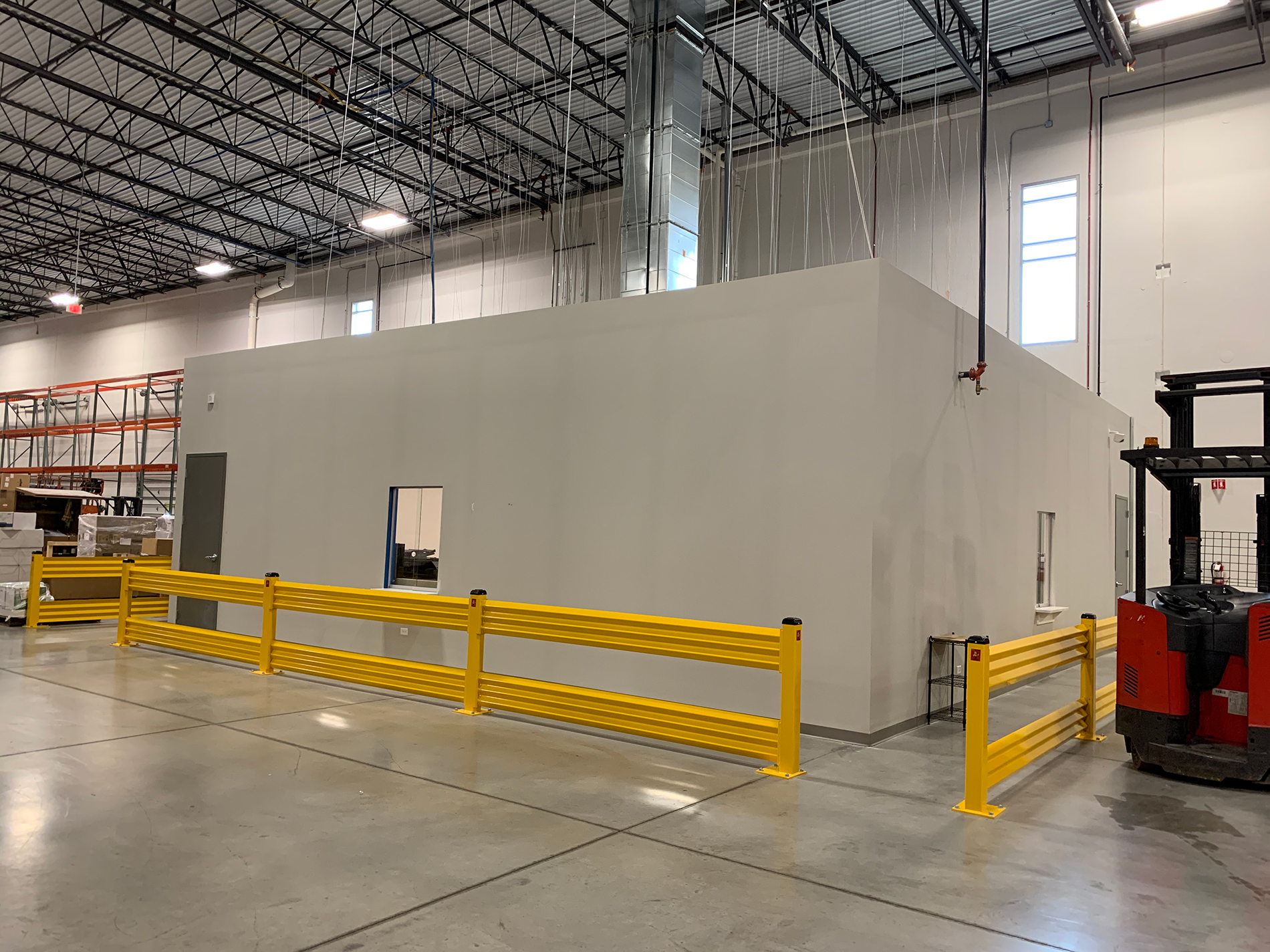 A large warehouse with shiny floors contains a small white room surrounded by yellow railings. A forklift sits to the right of the room.