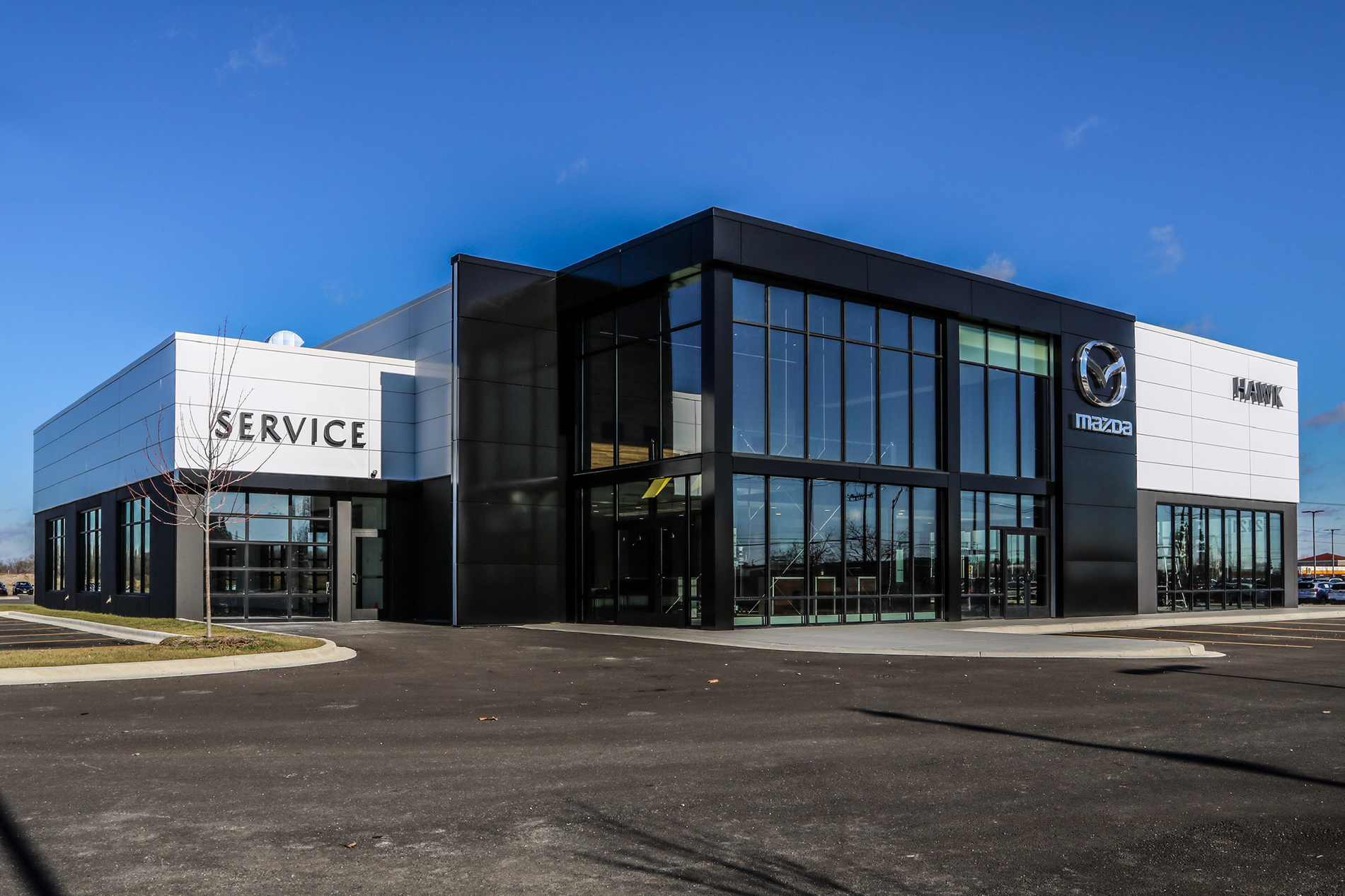 The Hawk Auto Dealership is a building with two stories of glass walls at the entrance and a side driveway with a sign that says “Service.” It was built by Keeley Construction, Inc.