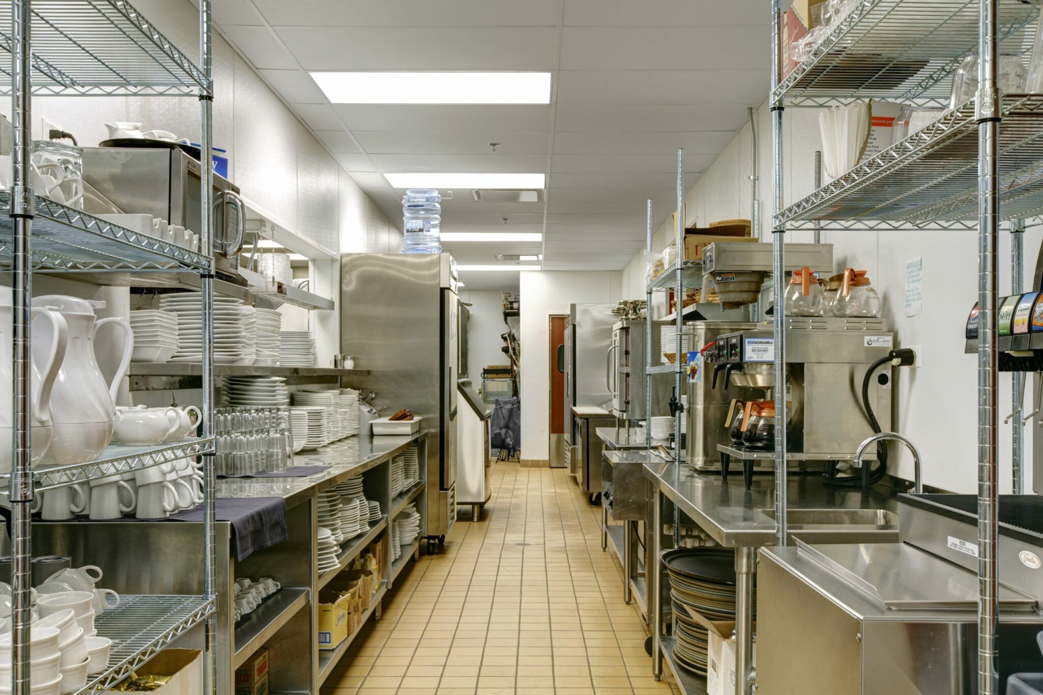 A view down the long walkway of a galley kitchen in Crazy Pour. Racks, coolers, an ice machine, and other equipment line the right and left walls.