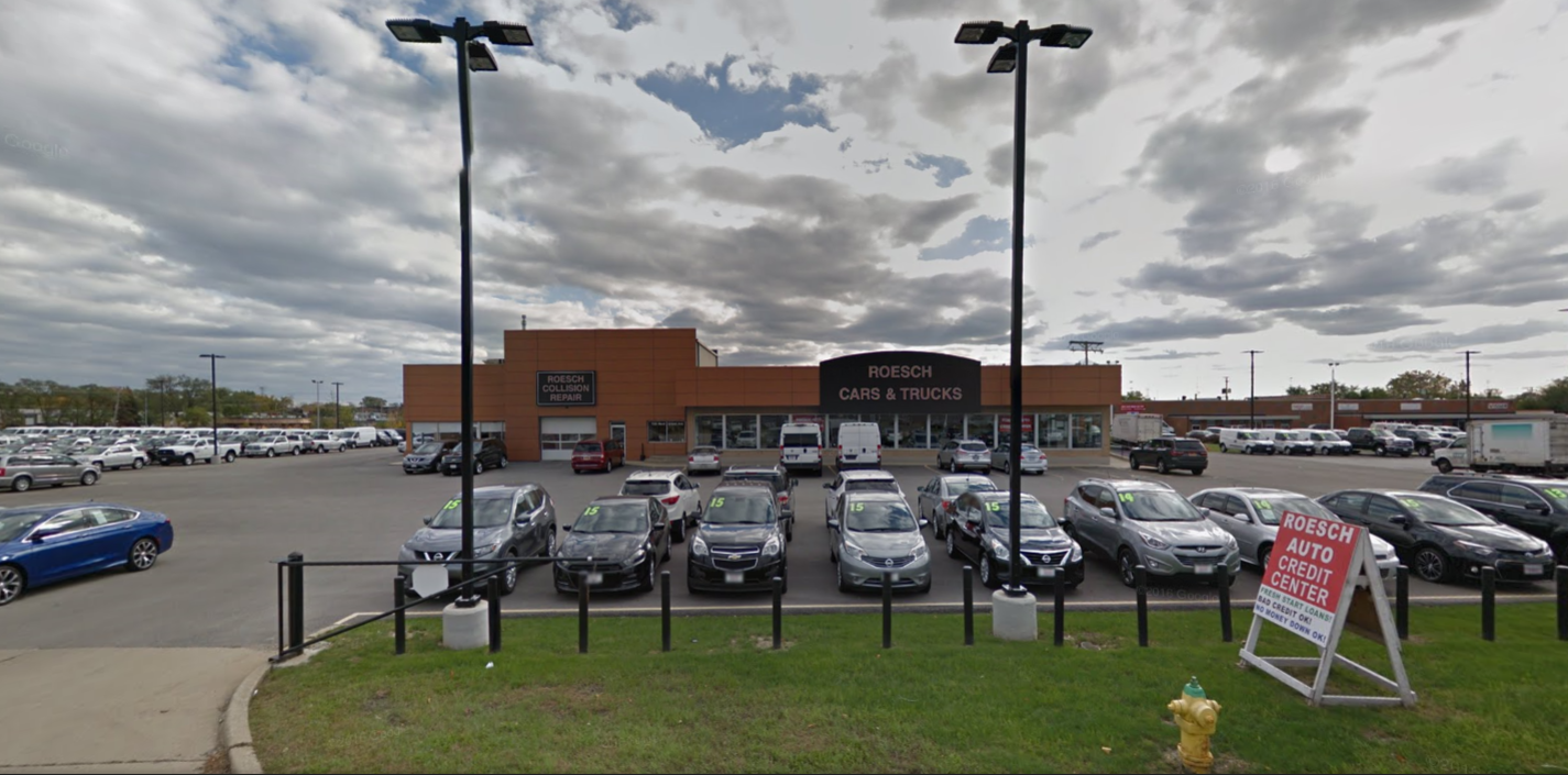 The parking lot and entrance to an auto dealership's showroom are behind a grass verge and two tall street lights.