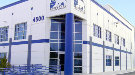 A view of the corner of a large blue and white facility, with mirrored windows at the entrance. The text on both corners of the building say, "Preferred Freezer Services."