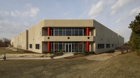 Two large red columns flank the flat corner entrance to the Ruprecht Meat Company Processing Facility. The large building stretches out behind the front doors to the left and right on a campus of manicured grass and trees.