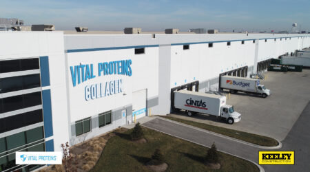 An aerial view of the outside of a long distribution area with docks and semi trucks. A large sign on the side says, "Vital Proteins Collagen."