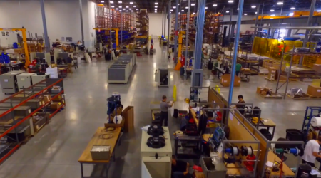 A second-floor view of a giant warehouse space, bustling with work, construction, and assembly amid tall metal shelves and workspaces.