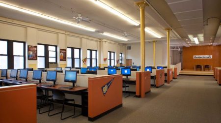 A classroom with short cubical walls divides the room into right and left-side computer workspaces facing the front dias. The wall in front has a banner that reads “Ombudsman.”